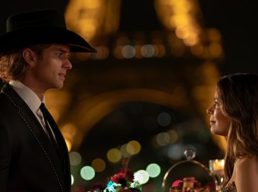 Miranda Cosgrove und Pierson Fode in The Wrong Paris: Ein Mann im Anzug und Cowboy-Hut steht in einem romantischen Moment einer Frau im Abendkleid unter dem beleuchteten Eiffelturm gegenüber.