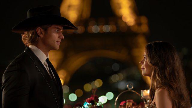 Miranda Cosgrove und Pierson Fode in The Wrong Paris: Ein Mann im Anzug und Cowboy-Hut steht in einem romantischen Moment einer Frau im Abendkleid unter dem beleuchteten Eiffelturm gegenüber.