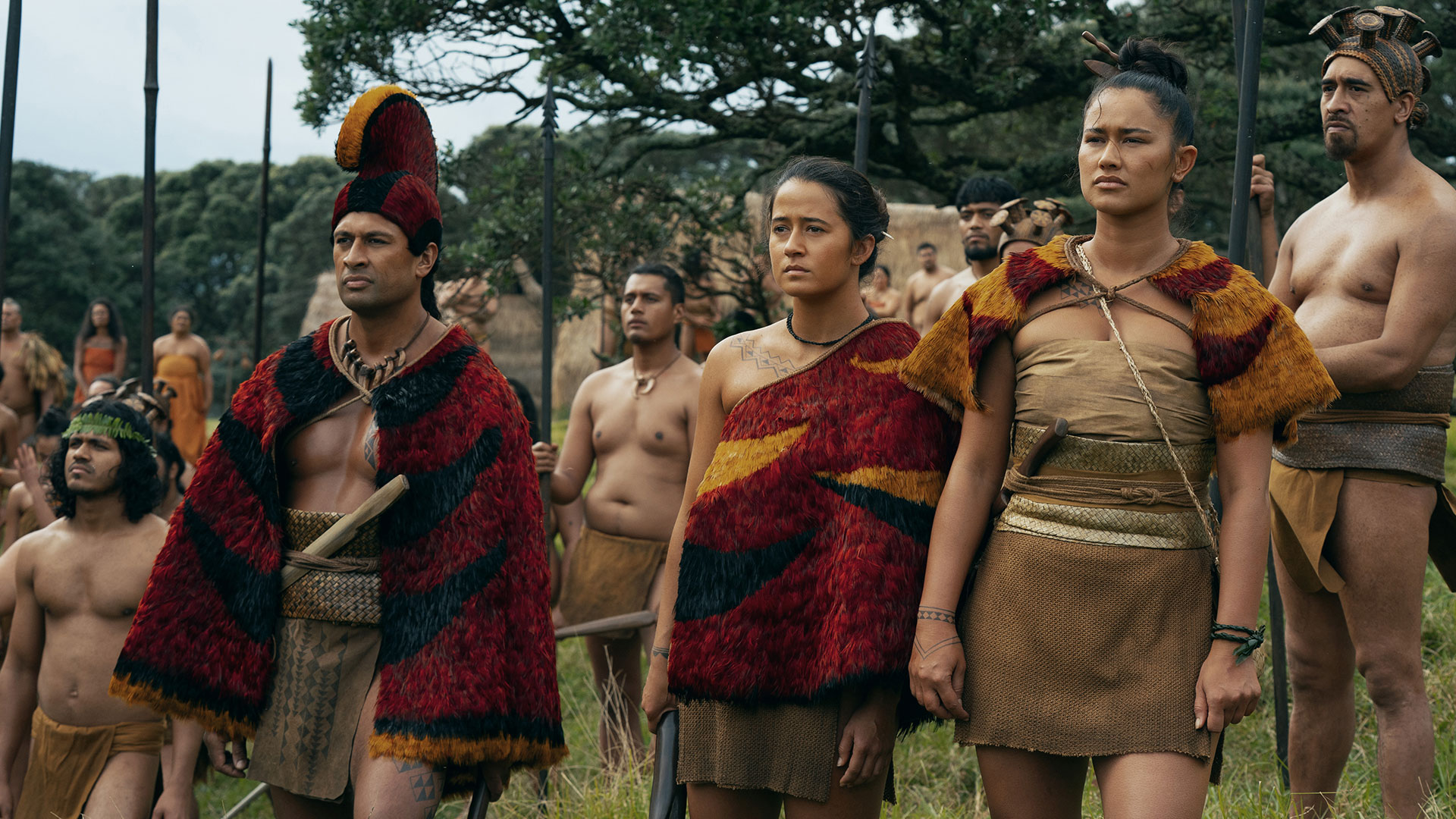 Auf dem Bild in dem Artikel zum „Chief of War“-Ende erklärt steht eine größere Gruppe Menschen in einer Wiese. Im Vordergrund sind drei Personen mit roten Umhängen zu sehen, dahinter stehen weitere mit Speeren.