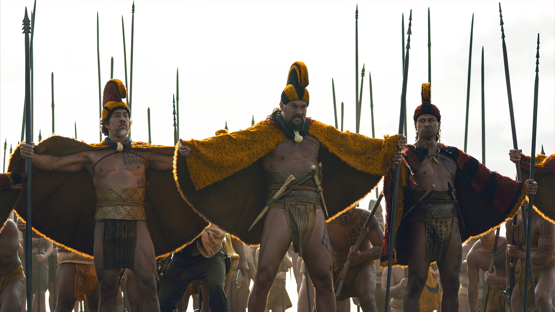 Auf dem Bild in dem Artikel zum „Chief of War“-Ende erklärt stehen drei Männer mit geöffneten Umhängen in Gelb und Rot vor einer Gruppe von Menschen mit Speeren. 