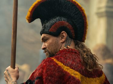 Auf dem Bild in dem Artikel zum „Chief of War“-Ende erklärt ist ein Mann mit rotem und gelben Umhang und aufwendigem Kopfschmuck zu sehen. Er hält einen Speer und schaut ernst zur Seite.