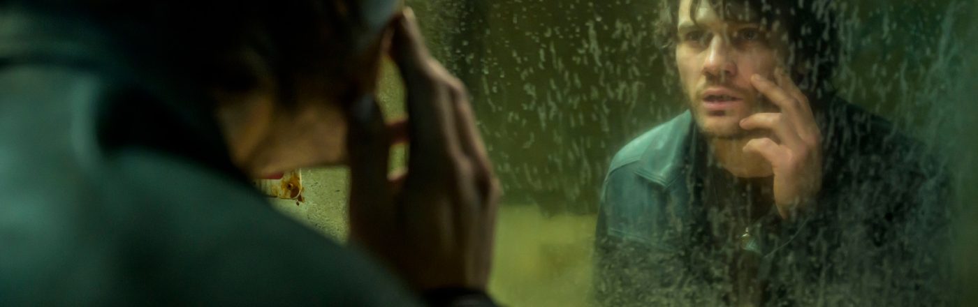 In dieser Szene aus den Spieleverfilmungen 2026 ist James Sunderland, gespielt von Jeremy Irvine, vor einem schmutzigen Spiegel zu sehen. Er betrachtet sich nachdenklich und leicht verstört, während er mit der Hand sein Gesicht berührt. Die düstere Stimmung und das heruntergekommene Badezimmer deuten auf psychische Belastung und innere Konflikte hin.