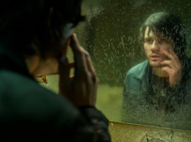 In dieser Szene aus den Spieleverfilmungen 2026 ist James Sunderland, gespielt von Jeremy Irvine, vor einem schmutzigen Spiegel zu sehen. Er betrachtet sich nachdenklich und leicht verstört, während er mit der Hand sein Gesicht berührt. Die düstere Stimmung und das heruntergekommene Badezimmer deuten auf psychische Belastung und innere Konflikte hin.