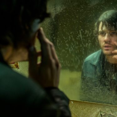 In dieser Szene aus den Spieleverfilmungen 2026 ist James Sunderland, gespielt von Jeremy Irvine, vor einem schmutzigen Spiegel zu sehen. Er betrachtet sich nachdenklich und leicht verstört, während er mit der Hand sein Gesicht berührt. Die düstere Stimmung und das heruntergekommene Badezimmer deuten auf psychische Belastung und innere Konflikte hin.