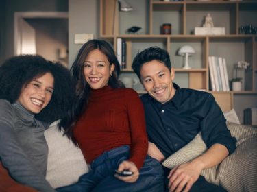 Vodafone TV Connect: Zwei Frauen und ein Mann sitzen auf einem Sofa. Eine Frau hat eine Fernbedienung in der Hand und sie schauen begeistert in die Kamera, wo der Fernsehr steht.