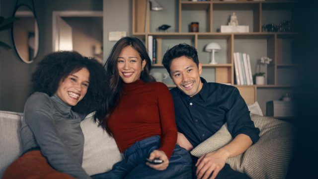 Vodafone TV Connect: Zwei Frauen und ein Mann sitzen auf einem Sofa. Eine Frau hat eine Fernbedienung in der Hand und sie schauen begeistert in die Kamera, wo der Fernsehr steht.