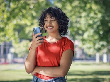 Plötzlich hoher Datenverbrauch: junge Frau schaut lächelnd auf ihr Handy in einem Park.
