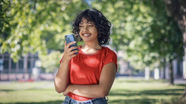 Plötzlich hoher Datenverbrauch: junge Frau schaut lächelnd auf ihr Handy in einem Park.