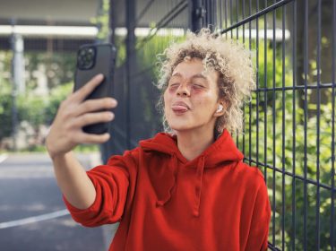 WhatsApp Datenverbrauch: Junge Frau steht draußer in urbaner Umgebung und streckt die Zunge in Richtung ihrer Handykamera heraus.