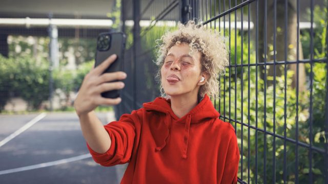 WhatsApp Datenverbrauch: Junge Frau steht draußer in urbaner Umgebung und streckt die Zunge in Richtung ihrer Handykamera heraus.