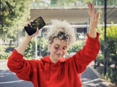 Datenverbrauch Social Media: Junge blonde Frau mit lockigen Haaren tanzt ausgelassen mit ihrem Handy in der Hand.