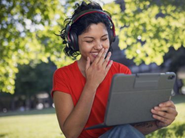Datenverbrauch Internetradio: Junge Frau sitzt mit Kopfhörern in einem Park und schaut lachend auf ihr Tablet