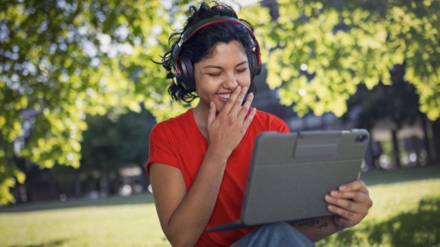 Datenverbrauch Internetradio: Junge Frau sitzt mit Kopfhörern in einem Park und schaut lachend auf ihr Tablet