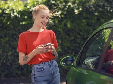 Roaming am iPhone aktivieren: Junge Frau mit ihrem Handy läuft an einem grünen Auto vorbei