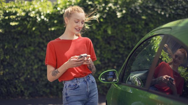 Roaming am iPhone aktivieren: Junge Frau mit ihrem Handy läuft an einem grünen Auto vorbei