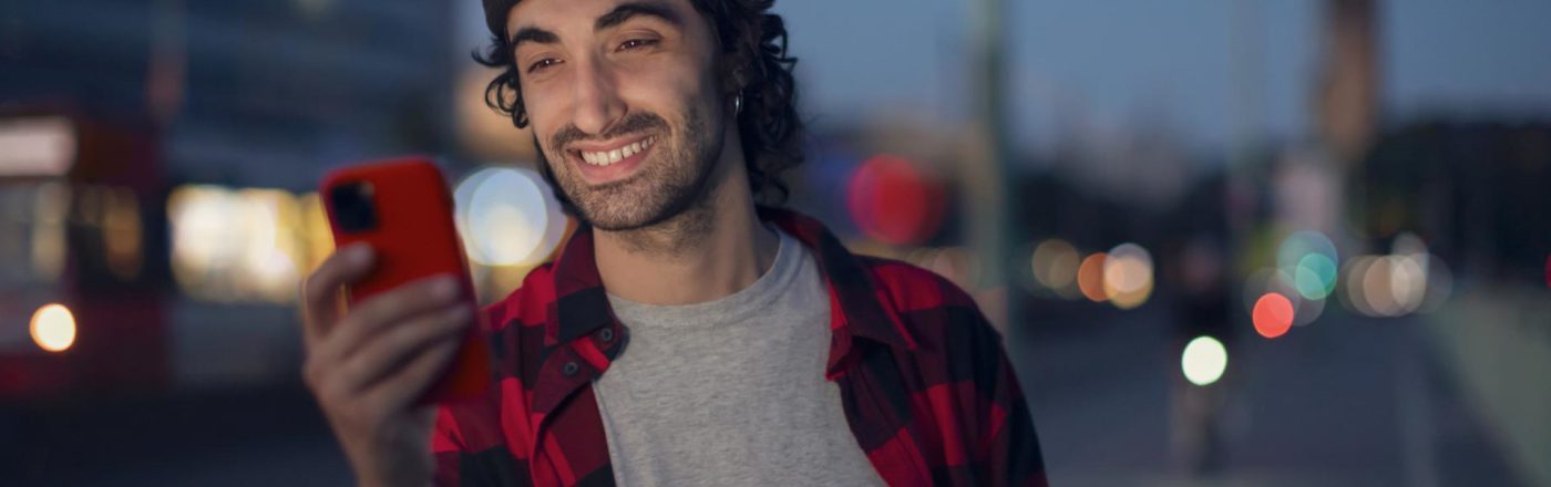 Roaming Android aktivieren: Mann in kariertem Hemd steht abends auf der Straße und schaut lächelnd auf sein Handy