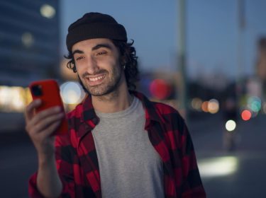 Roaming Android aktivieren: Mann in kariertem Hemd steht abends auf der Straße und schaut lächelnd auf sein Handy