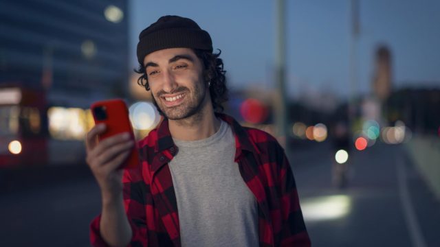 Roaming Android aktivieren: Mann in kariertem Hemd steht abends auf der Straße und schaut lächelnd auf sein Handy