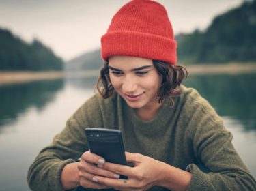 Roaming Samsung: Junge Frau sitzt mit einer roten Mütze und ihrem Handy in der Hand an einem See