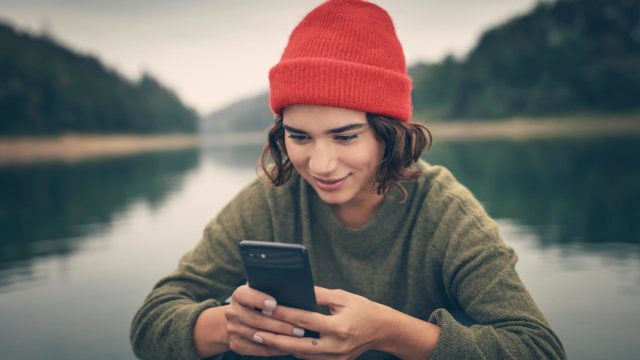 Roaming Samsung: Junge Frau sitzt mit einer roten Mütze und ihrem Handy in der Hand an einem See