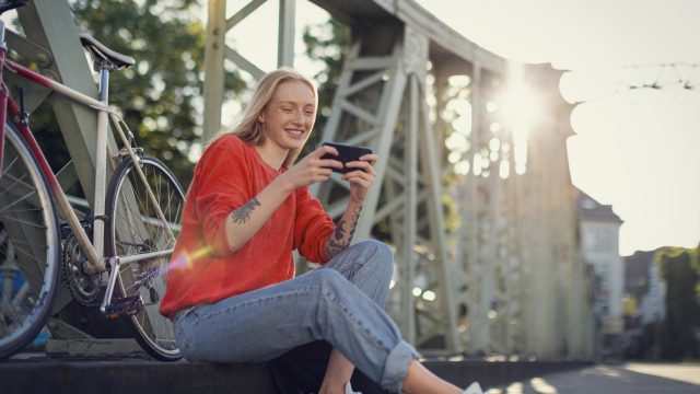 Roaming Xiaomi funktioniert nicht: junge Frau sitzt auf einer Brücke lächelnd neben ihrem Farrahd und schaut auf ihr Handy