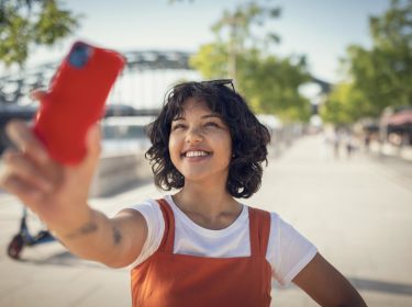 Roaming Gebühren EU: Junge Frau in rotem Kleid steht auf belebter Promenade und macht ein Selfie von sich