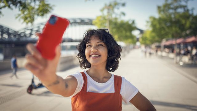 Roaming Gebühren EU: Junge Frau in rotem Kleid steht auf belebter Promenade und macht ein Selfie von sich