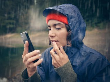 Kein Internet trotz Roaming: Junge Frau in Regemantel benutzt im Regen ihr Handy