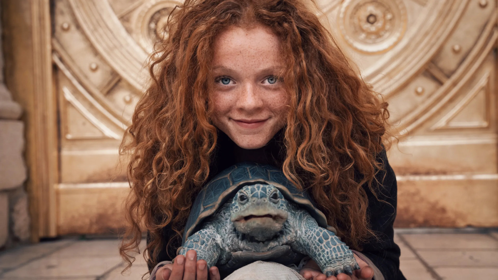 Alexa Goodall in Momo: Ein junges Mädchen mit roten Locken und strahlend blauen Augen lächelt verschmitzt. Unter ihrem Gesicht eine blaue Schildkröte.