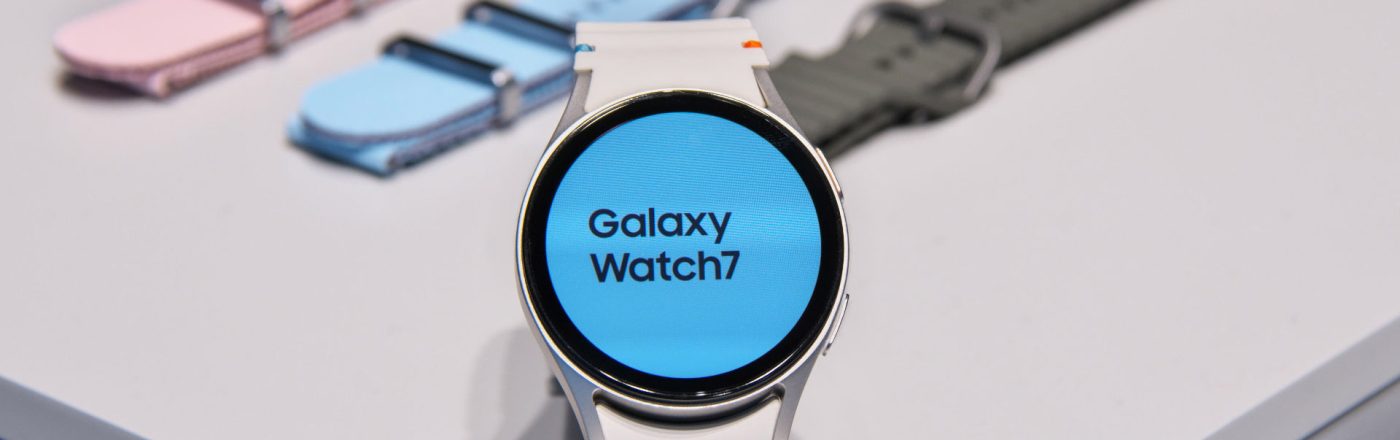 Die Galaxy Watch7 aufgestellt auf einem hellen Tisch, dahinter farbige Armbänder.