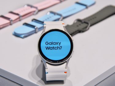 Die Galaxy Watch7 aufgestellt auf einem hellen Tisch, dahinter farbige Armbänder.
