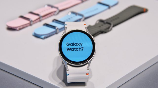 Die Galaxy Watch7 aufgestellt auf einem hellen Tisch, dahinter farbige Armbänder.