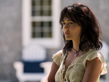 Auf dem Bild zur Serie Solange wir lügen Staffel 2 ist Emily Alyn Lind in ihrer Rolle als Cadence Sinclair Eastman zu sehen. Sie steht leicht seitlich im Bild, mit ernster Miene und nachdenklichem Blick. Ihr cremefarbenes Kleid mit Rüschen und das dunkle, leicht zerzauste Haar betonen den dramatischen Ton der Szene. Der Hintergrund zeigt unscharfe Gartenmöbel vor einer Hausfassade.