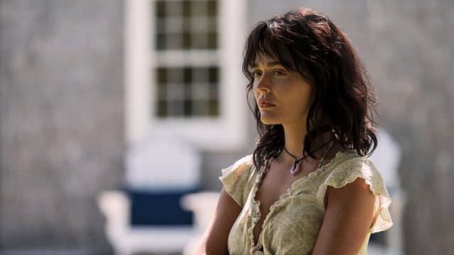 Auf dem Bild zur Serie Solange wir lügen Staffel 2 ist Emily Alyn Lind in ihrer Rolle als Cadence Sinclair Eastman zu sehen. Sie steht leicht seitlich im Bild, mit ernster Miene und nachdenklichem Blick. Ihr cremefarbenes Kleid mit Rüschen und das dunkle, leicht zerzauste Haar betonen den dramatischen Ton der Szene. Der Hintergrund zeigt unscharfe Gartenmöbel vor einer Hausfassade.