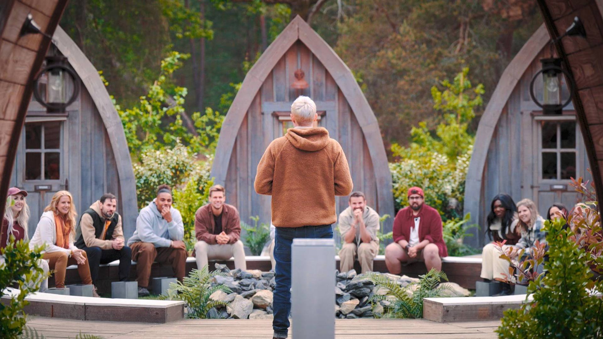 In den GigaTV-Highlights im Oktober 2025 ist eine Szene aus der Reality-Show „Werwölfe – Das Spiel von List und Täuschung“ zu sehen. Eine Gruppe von Teilnehmer:innen sitzt im Halbkreis im Außenbereich vor kleinen Holzhütten. Eine Person mit brauner Fleecejacke steht mit dem Rücken zur Kamera und spricht die Gruppe an.