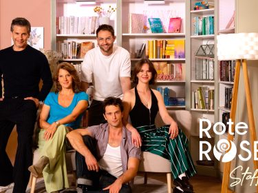 Auf dem Bild zur Rote Rosen Staffel 24 posieren fünf Schauspieler:innen vor einem hellen Bücherregal. Zwei Personen sitzen auf einem Sofa, drei stehen oder sitzen locker drumherum.