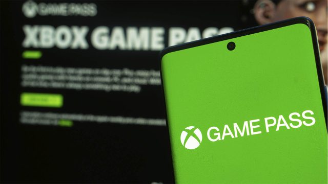 Ein Smartphone zeigt das Logo des Xbox Game Pass; im Hintergrund ist die geöffnete Game-Pass-App unscharf zu erkennen.