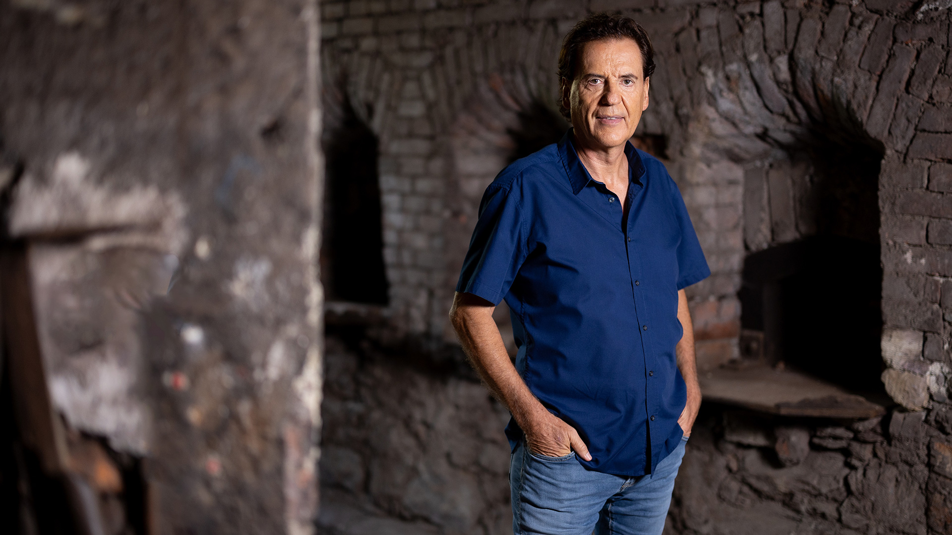 Jörg Dahlmann steht auf dem Bild zu „Das große Promi-Büßen“ 2025 mit blauem Hemd und Jeans in einem rustikalen Raum mit groben Steinwänden.