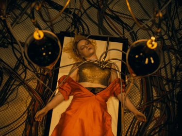 In dieser Szene aus The Bride 2026 liegt Jessie Buckley als die Braut bewusstlos auf einem Tisch, umgeben von zahlreichen Schläuchen und Kabeln. Metallene Vorrichtungen sind an ihrer Brust befestigt, während ihr rotes Kleid dramatisch inszeniert ist. Die düstere, futuristische Atmosphäre vermittelt ein Gefühl von Kontrollverlust und technischer Manipulation.