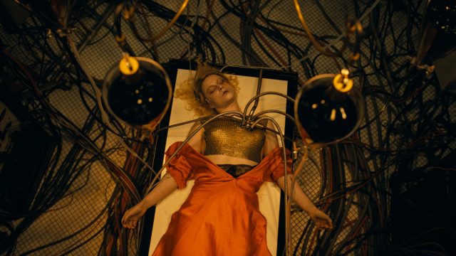 In dieser Szene aus The Bride 2026 liegt Jessie Buckley als die Braut bewusstlos auf einem Tisch, umgeben von zahlreichen Schläuchen und Kabeln. Metallene Vorrichtungen sind an ihrer Brust befestigt, während ihr rotes Kleid dramatisch inszeniert ist. Die düstere, futuristische Atmosphäre vermittelt ein Gefühl von Kontrollverlust und technischer Manipulation.