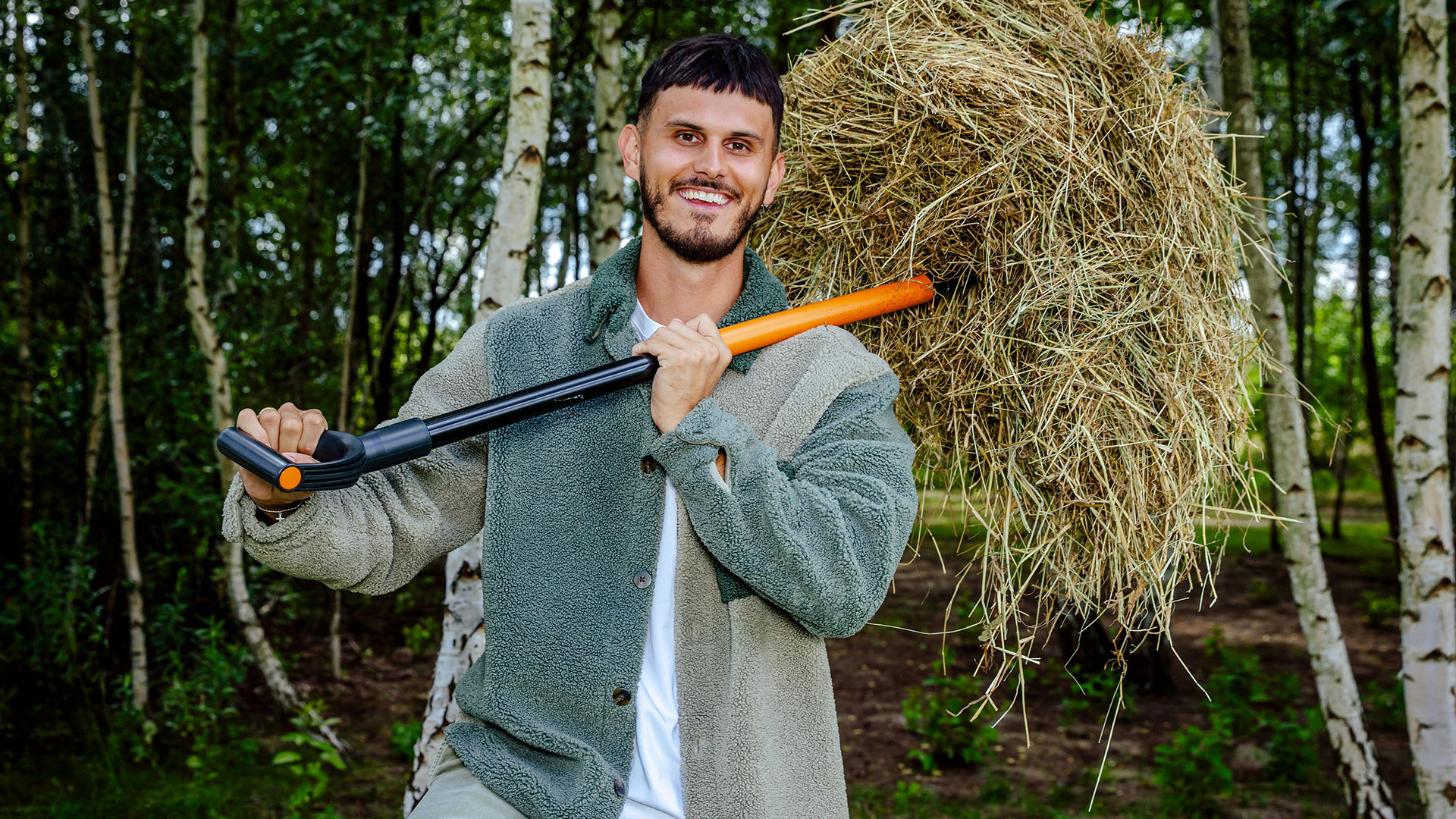 Auf dem Bild zur Sendung Der Promihof ist Nino Sifkovits zu sehen, der als Aufpasser auf dem Hof arbeitet. Er steht lachend in einem Waldstück und trägt ein Bündel Heu auf einer modernen Mistgabel über der Schulter. Seine Kleidung wirkt robust und ländlich, passend zur Umgebung. Die Stimmung ist entspannt und naturverbunden.