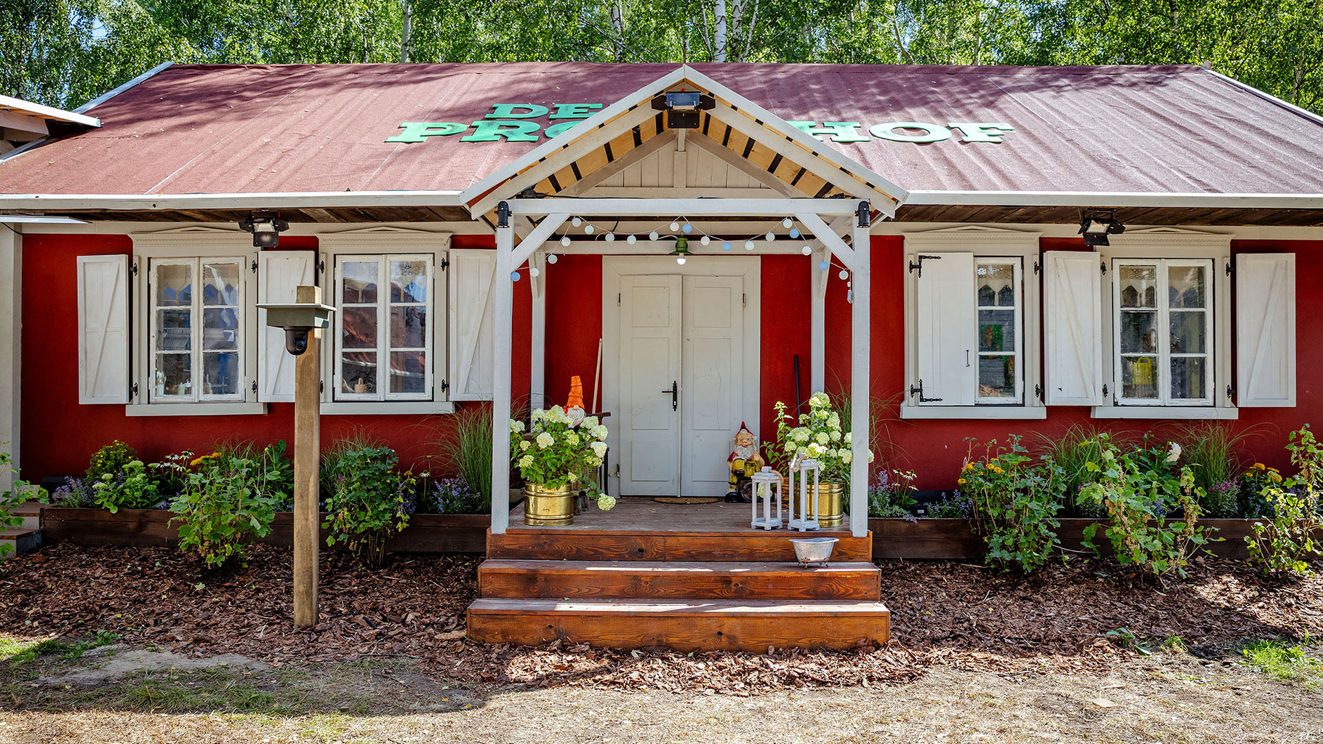 Auf diesem Bild zur Reality-Show Der Promihof ist der rot gestrichene Bauernhof mit weißem Giebeldach und dekorativem Eingangsbereich zu sehen. Vor der zentralen Doppeltür stehen Blumen, Gartenfiguren und Laternen, was eine einladende Atmosphäre schafft. Das Schild „Der Promihof“ ist groß auf dem Dach angebracht und signalisiert den zentralen Drehort der Show.