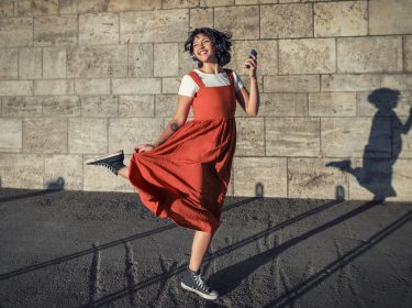 Datenverbrauch am iPhone: Junge Frau in rotem Kleid tanzt ausgelassen mit ihrem Smartphone in der Sonne vor einer Wand.