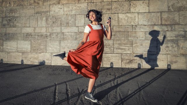 Datenverbrauch am iPhone: Junge Frau in rotem Kleid tanzt ausgelassen mit ihrem Smartphone in der Sonne vor einer Wand.