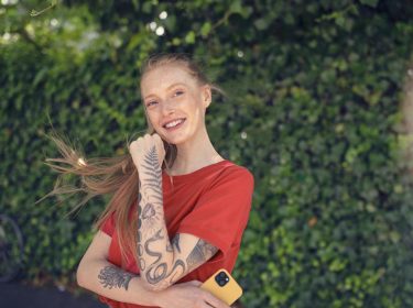 Android Datenverbrauch: Junge Frau in rotem T-Shirt steht mit ihrem Smartphone vor einer Hecke.