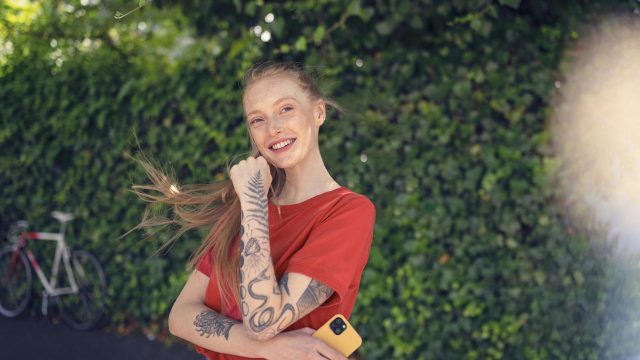 Android Datenverbrauch: Junge Frau in rotem T-Shirt steht mit ihrem Smartphone vor einer Hecke.