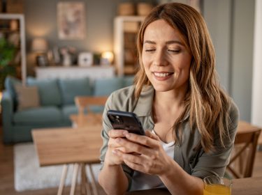 Im Symbolbild für den Handy Garantieverlängerung-Ratgeber sitzt eine lächelnde Frau entspannt in einem modern eingerichteten Wohnzimmer und schaut auf ihr Smartphone.