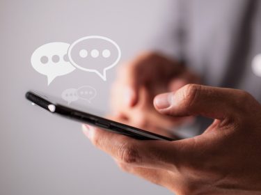 Eine Person hält ein Smartphone in den Händen, Sprachblasen symbolisieren Kommunikation via Messenger.