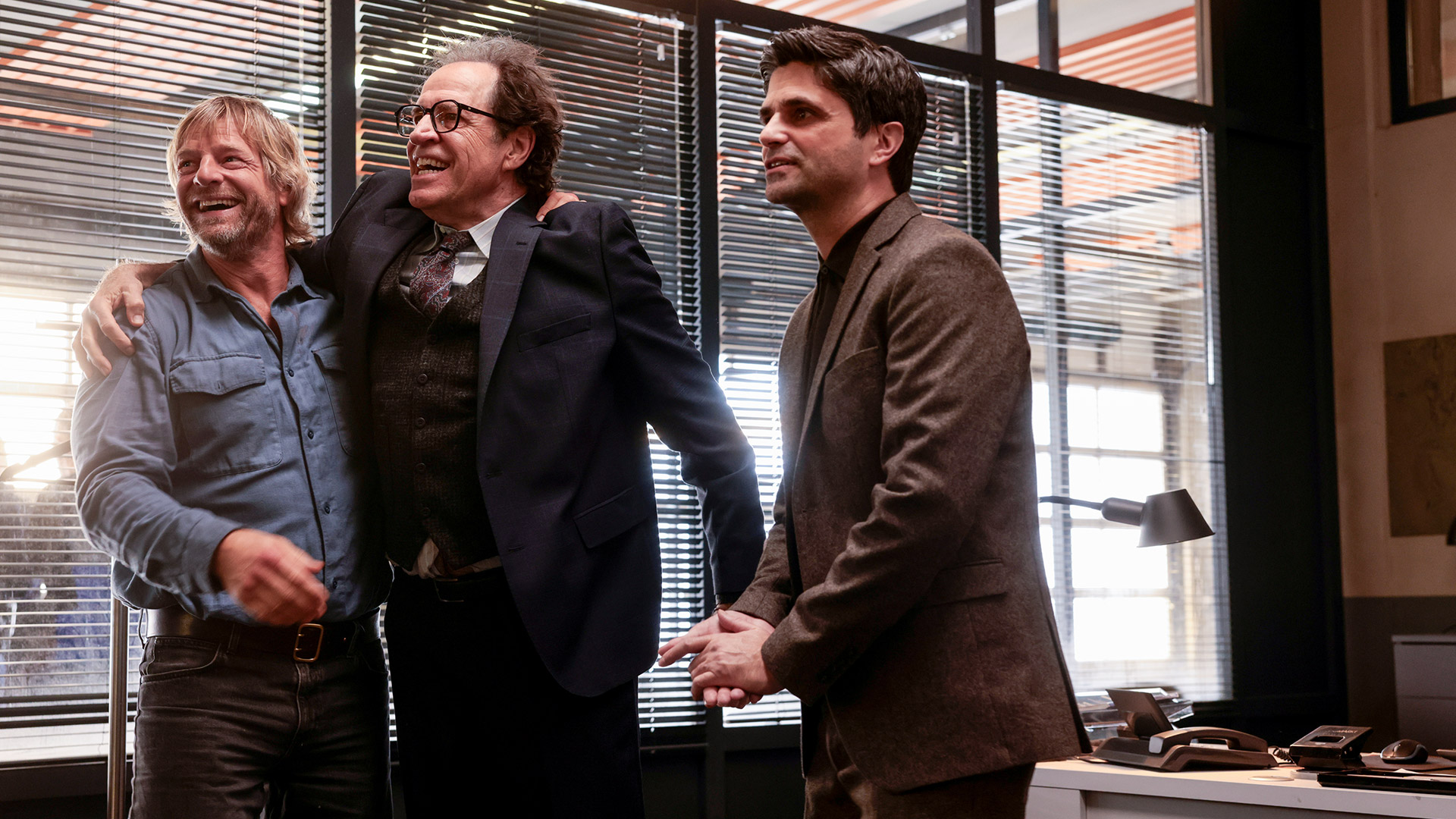 In einer Szene aus Der letzte Bulle stehen drei Männer in einem Büro. Links lacht Mick Brisgau, gespielt von Henning Baum, in einem blauen Hemd. In der Mitte ein fröhlicher Mann im Anzug, rechts ein ernsterer Kollege.