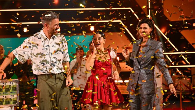 Dieses Bild zu den Promi Big Brother Gewinner:innen zeigt die frisch gekürte Siegerin Leyla Lahouar in einem roten Kleid, umgeben von den Moderatoren Jochen Schropp und Marlene Lufen. Um sie regnet es goldenes Konfetti, während sie die Trophäe in der Hand hält und überwältigt lächelt. Im Hintergrund jubelt das Publikum – ein Moment voller Emotionen und Showglanz.